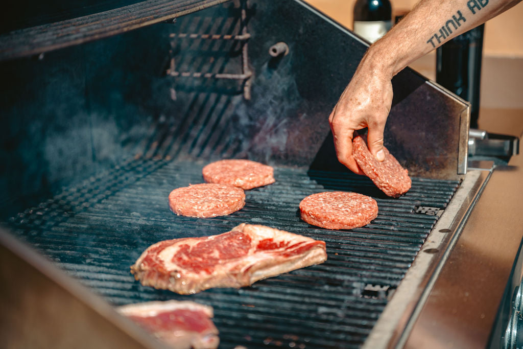 Angus beef steaks and burger patties grilling over open flame, pasture-raised and dry-aged for rich, bold flavor.