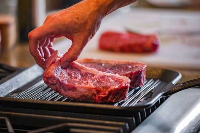 Two pasture-raised KC strip steaks searing in a hot pan, dry-aged for bold, beefy flavor.